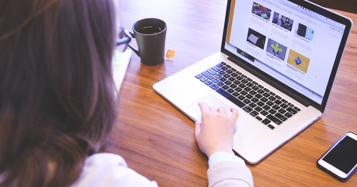 A person using a laptop on a wooden table browses a digital marketing website filled with images. A cup of coffee and a smartphone are nearby, while they navigate the site using the trackpad with their right hand.