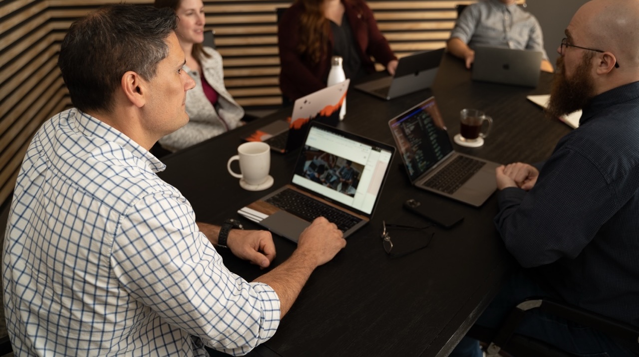 People sitting around a conference table with open laptops, engaged in a lively discussion about design. One person is wearing a checkered shirt, another has a beard. Coffee mugs and glasses are scattered across the table, adding to the creative atmosphere of this office meeting. People sitting around a conference table with open laptops, engaged in a lively discussion about design. One person is wearing a checkered shirt, another has a beard. Coffee mugs and glasses are scattered across the table, adding to the creative atmosphere of this office meeting.