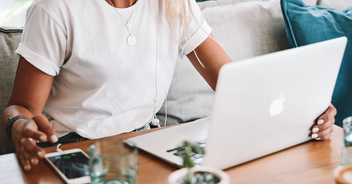 A person wearing a white t-shirt sits at a wooden table using a laptop for marketing tasks, with a smartphone in one hand. Earphones dangle from their neck, and a glass of water and small plant are nearby.