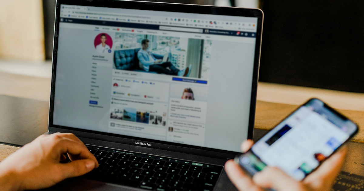A person using a laptop to browse a social media profile page for marketing purposes, while holding a smartphone in their other hand, seated at a desk with both screens visible.