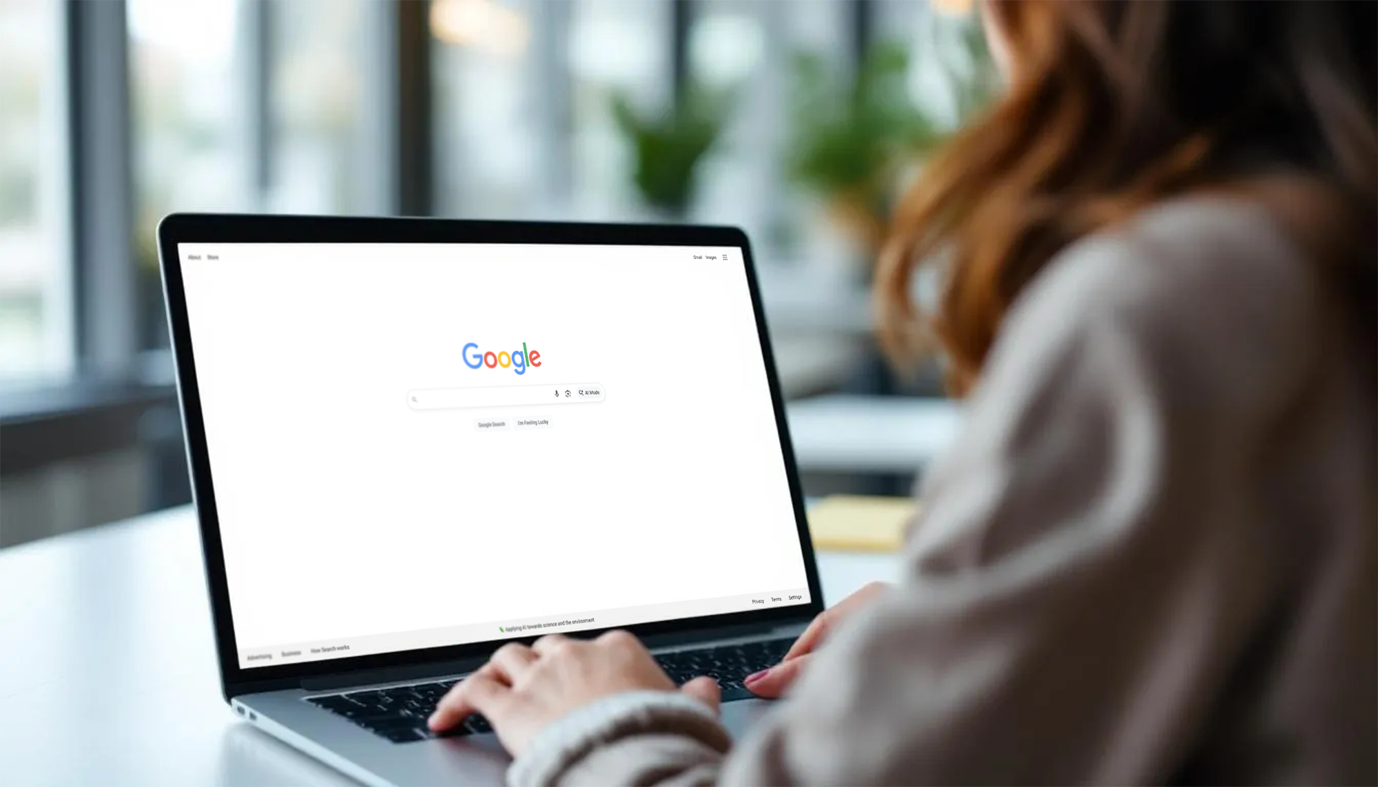 A person with long hair uses a laptop at a desk, the Google search homepage displayed as they research marketing strategies. The background is softly blurred with green plants and bright window light.