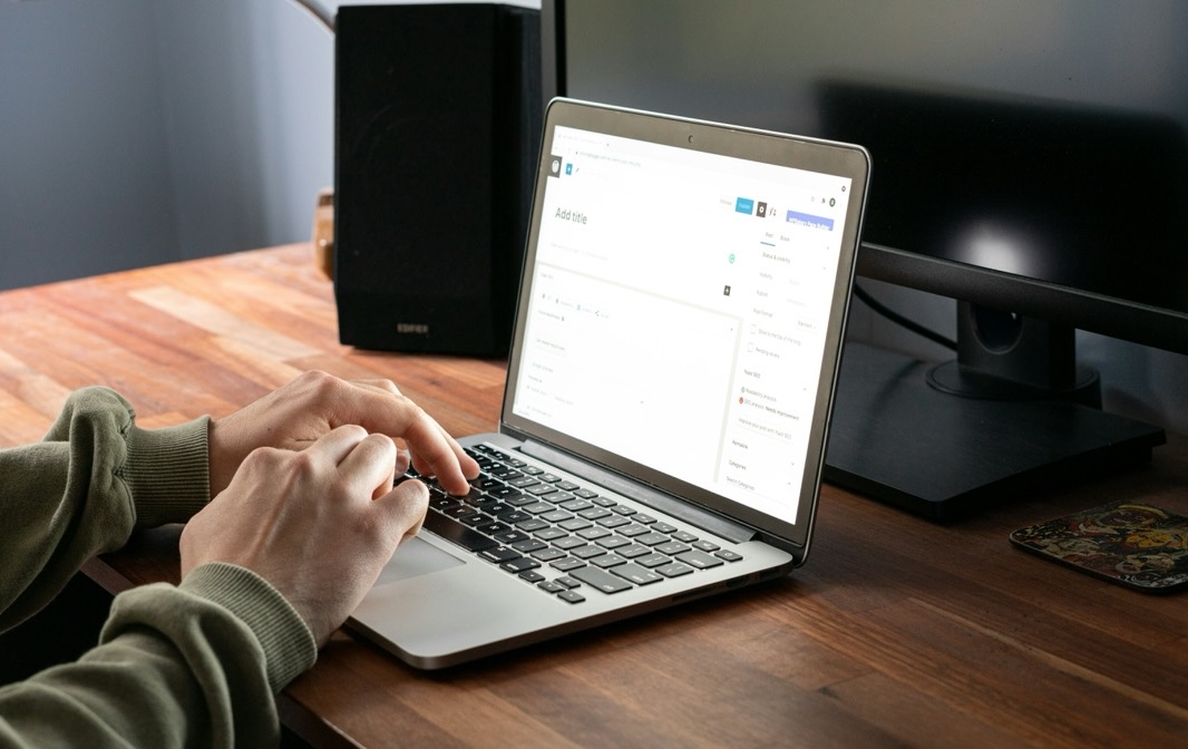 A person types on a laptop placed on a wooden desk next to a monitor, speaker, and mousepad, with a website content management interface displayed on the laptop screen.