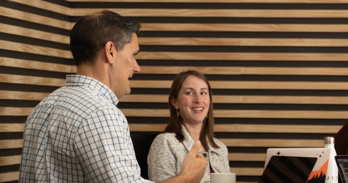 Two people sit at a table in an office with striped wooden walls. The man in the foreground gestures as he discusses marketing; the woman across from him smiles and listens. Laptops and a coffee mug are on the table.