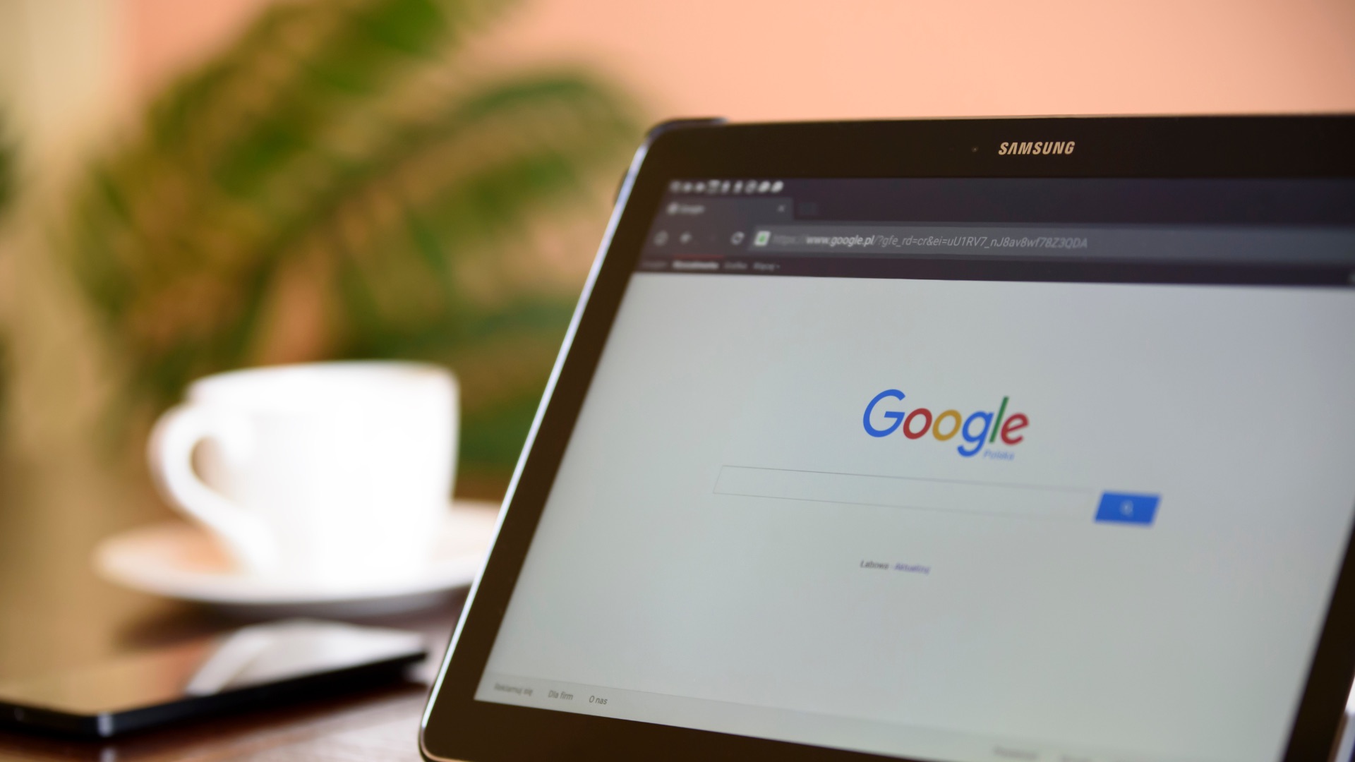 Why Your Google Ads Are Getting Clicks But No Conversions A Samsung tablet displays the Google homepage. In the blurred background, a white coffee cup and saucer, a smartphone with Google open on its screen, and a green plant are visible on a wooden surface.